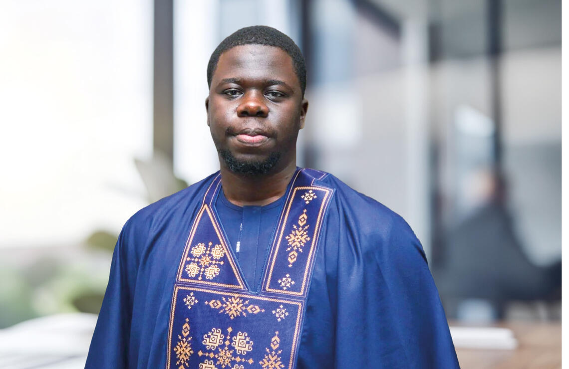 Une personne porte une tenue traditionnelle bleue ornée de motifs dorés et pose dans un environnement intérieur flou. DR
